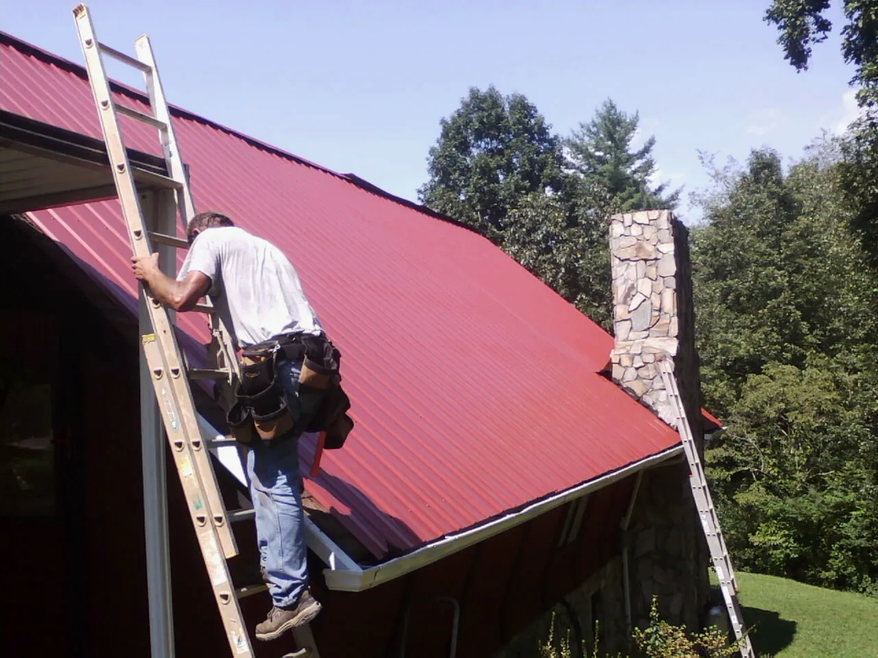 Roofer on a ladder beside a red metal roof and stone chimney — Soffit & Fascia Repair by Roofing Brothers in Mc Cutchenville